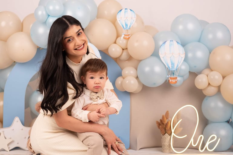 Mujer de cabello oscuro y niño con camisa clara en una decoración festiva con globos azules y beige.