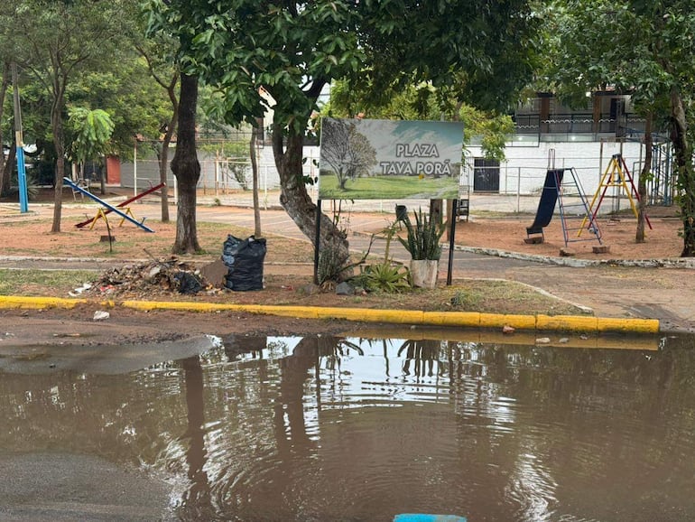 Letrero de 'Plaza Tava Porá' en área descuidada con juegos infantiles y basura acumulada.
