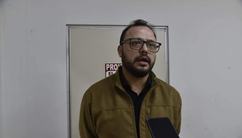 Hombre con barba, chaqueta marrón y gafas, en ambiente interior con pared blanca y puerta de fondo.
