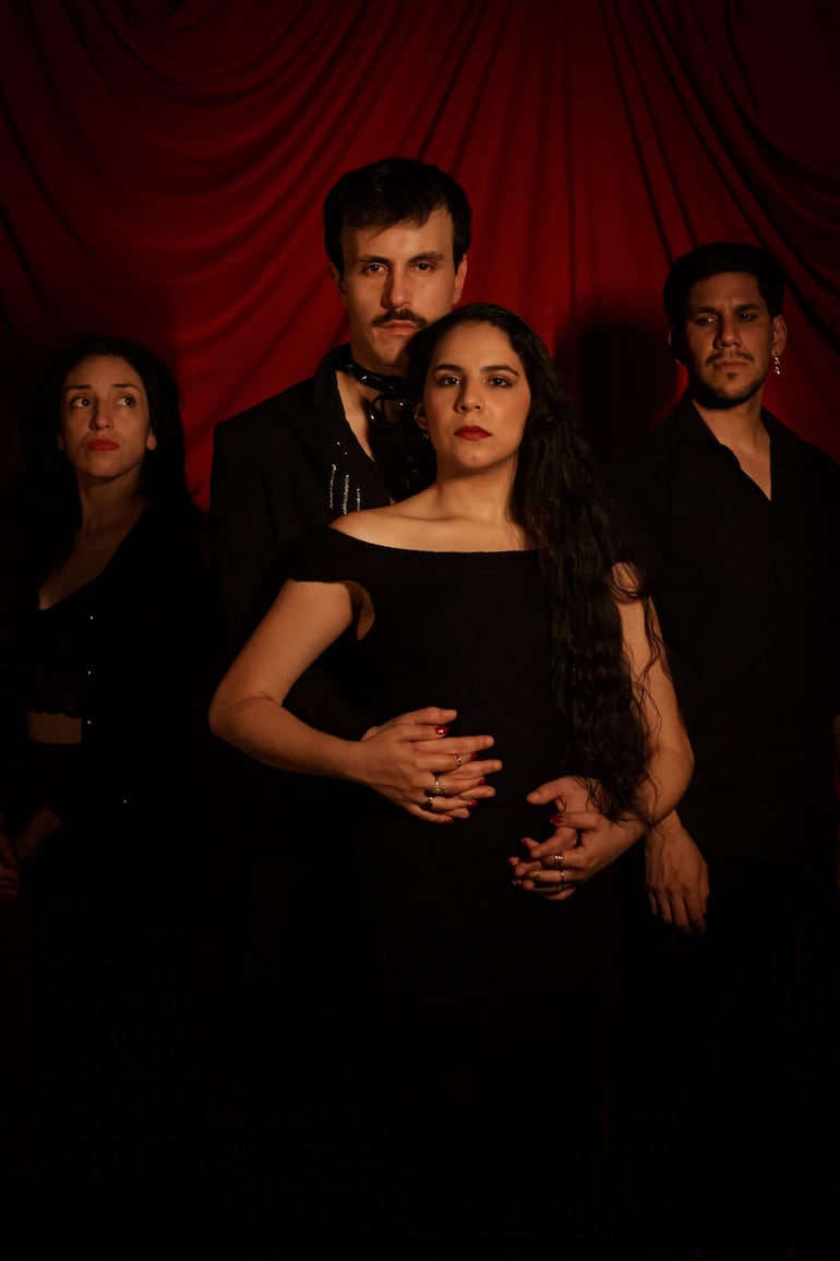 Cuatro miembros del elenco, dos mujeres con vestidos oscuros y dos hombres con atuendos serios, en un fondo de tela roja.