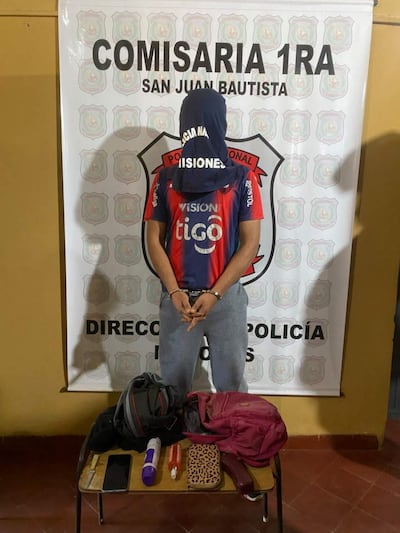 Hombre con camiseta roja y azul cubriendo su rostro, de pie en comisaría con manos cruzadas y objetos personales sobre la mesa.