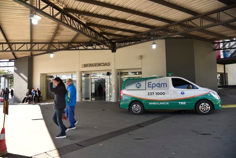 Mujer con teléfono y hombre con gorro azul aguardan en la entrada del hospital, junto a una ambulancia identificada.