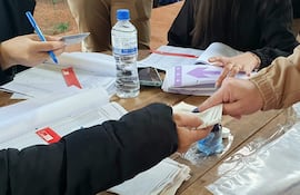 Personas en un centro de votación, con documentos y agua mineral en la mesa. Ambiente activo y de intercambio.