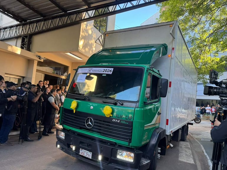 Un camión verde y blanco con el texto 'ELECCIONES MUNICIPALES 2026' estacionado, rodeado de periodistas en ambiente formal.