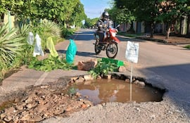 Calle con un bache profundo y agua acumulada, coches estacionados a los lados y vegetación en un día soleado.