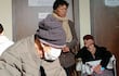 Hombre mayor con gorro y máscara facial escribe, mientras mujer de pie observa y otra mujer en silla de ruedas se cubre el rostro.