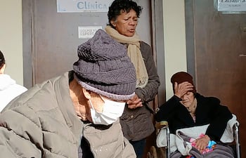 Hombre mayor con gorro y máscara facial escribe, mientras mujer de pie observa y otra mujer en silla de ruedas se cubre el rostro.