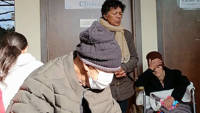 Hombre mayor con gorro y máscara facial escribe, mientras mujer de pie observa y otra mujer en silla de ruedas se cubre el rostro.
