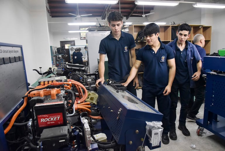 Cuatro jóvenes en camiseta polo oscuro observan un motor eléctrico en un taller educativo, atentos a la explicación de uno de ellos.