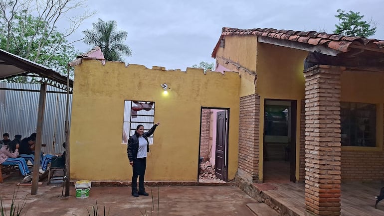 Mujer en camisa blanca y chaqueta negra señala daños en la pared de un edificio amarillo, con personas sentadas detrás.