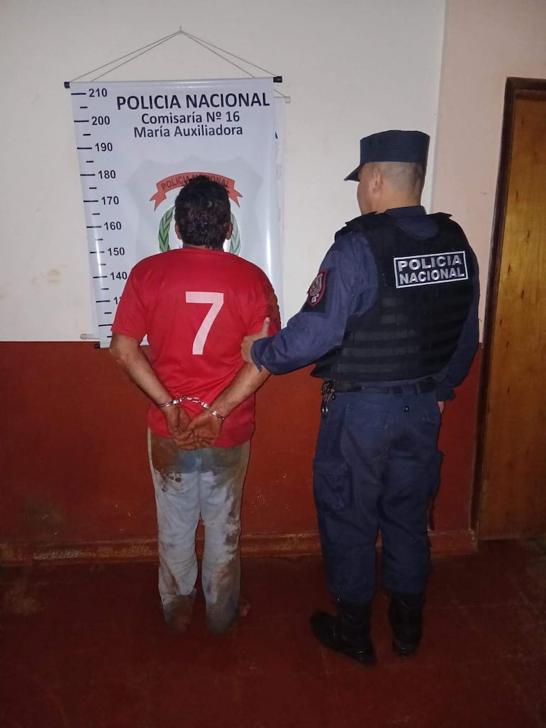 Hombre esposado con camiseta roja número 7, mientras un policía lo sostiene por el brazo en la comisaría con cartel de 'Policía Nacional'.