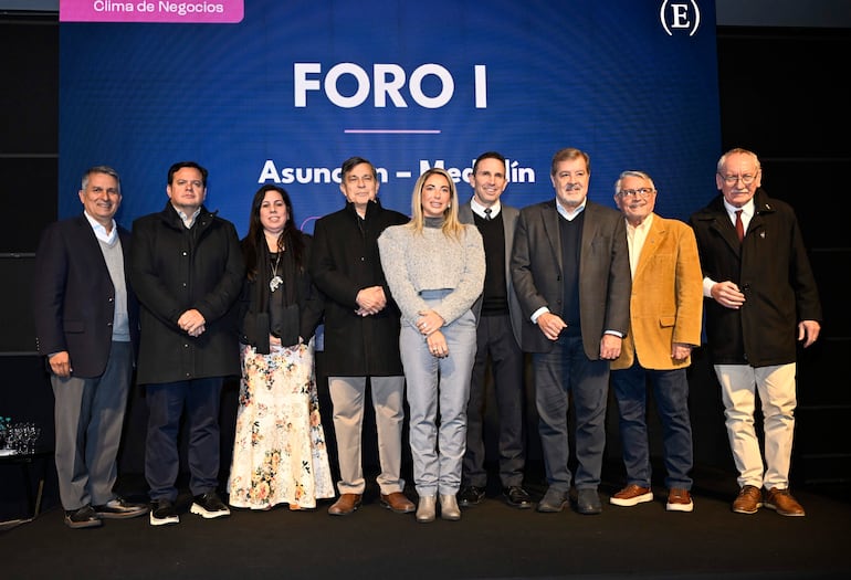 Ocho personas de pie en el escenario, hombres con trajes, mujeres con vestidos y conjuntos claros, fondo con diseño simple del foro.
