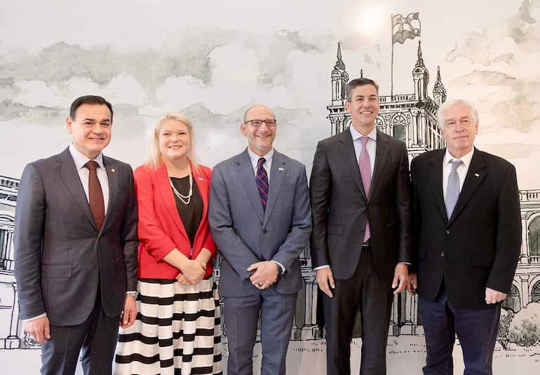Cinco individuos sonrientes, vestidos con trajes oscuros y coloridos, posan en un entorno oficial con un fondo ilustrado.