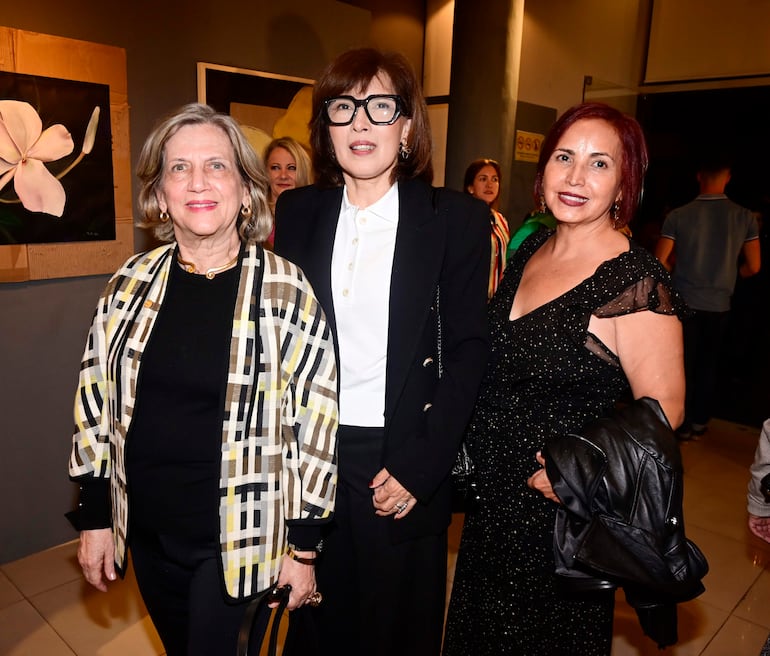 Tres mujeres sonrientemente conversan; una lleva abrigo de cuadros, otra camisa blanca y abrigo negro, y la última blusa negra con transparencias.