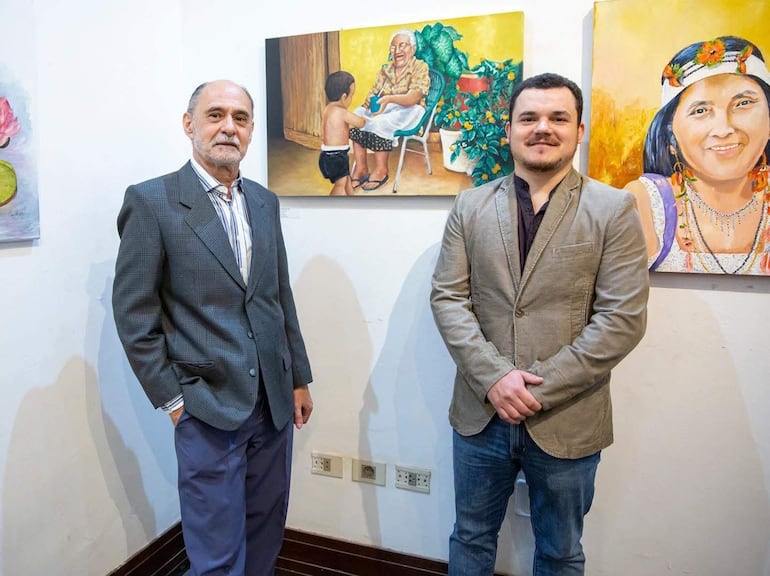 Hombre canoso con traje gris y otro con chaqueta clara posan juntos frente a pinturas en una galería de arte.