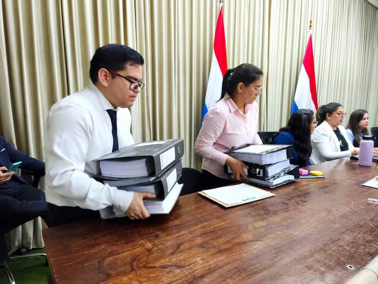 Hombre con gafas y corbata blanca, mujer en camisa rosa y tres mujeres sentadas, en un entorno formal con banderas paraguayas.