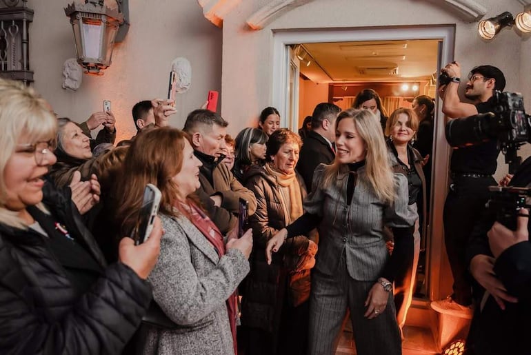 Mujer sonriente en primer plano, saludando a la audiencia, con un grupo de personas observando y capturando el momento con celulares.