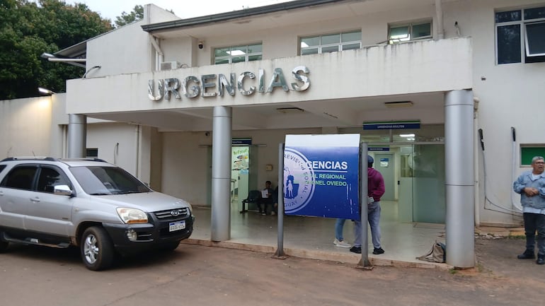 Personas en la entrada de un centro de salud, esperando atención, con un auto gris estacionado y cartel de 'URGENCIAS'.