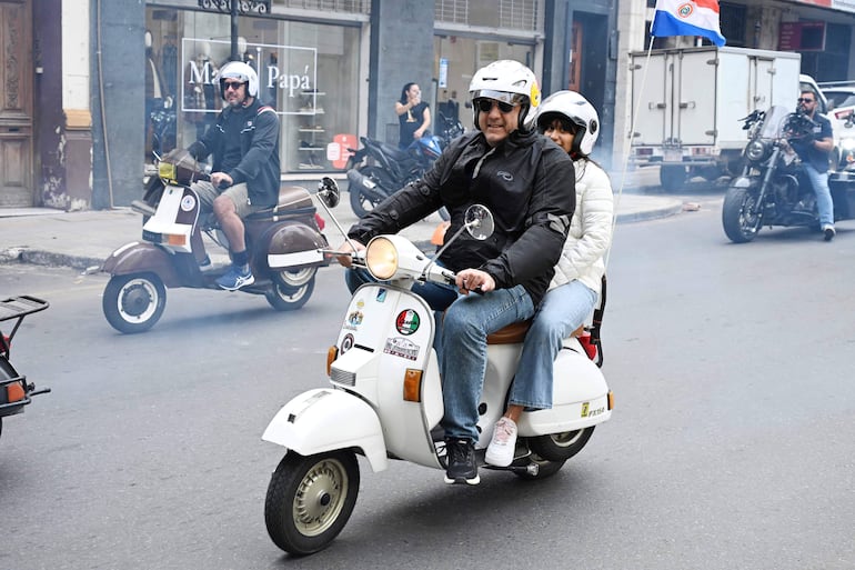 Un hombre con chaqueta negra y gafas conduce un scooter blanco, acompañado por una mujer con chaqueta blanca.