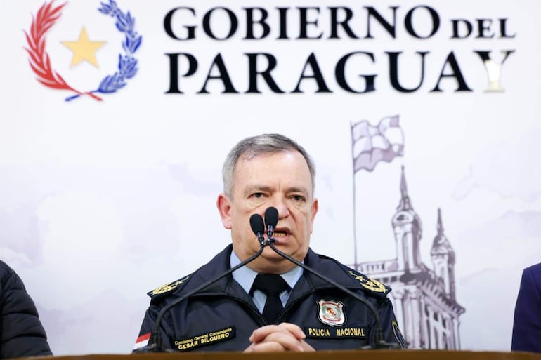 César Silguero en uniforme policial, hablando ante un micrófono en una conferencia llena de asistentes, con fondo gubernamental.
