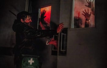 Hombre en abrigo oscuro, con mochila de primeros auxilios, presionando una puerta cerrada mientras se ven manos en las ventanas.