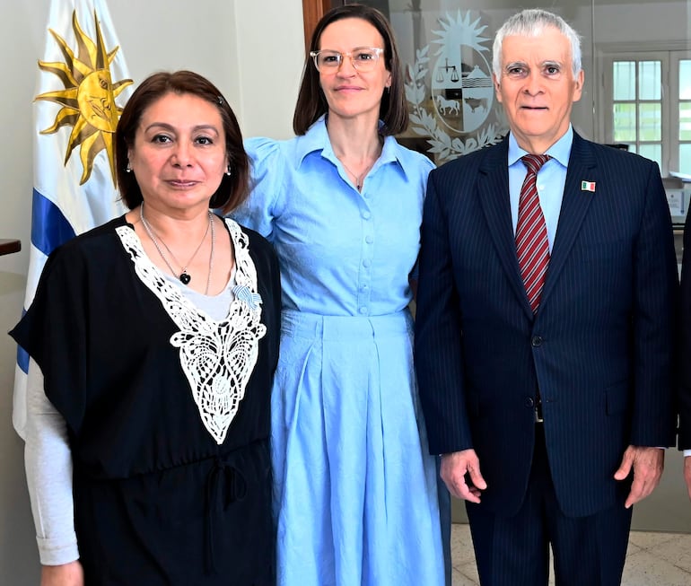 Mujer en blusa negra y mujer en blusa azul claro, junto a hombre en traje a rayas y corbata roja, posando en una reunión formal.