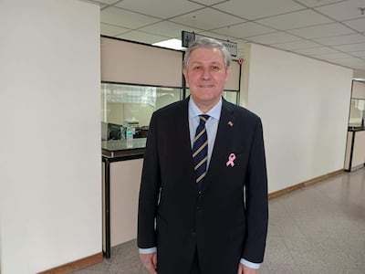 Hombre sonriente en traje oscuro y corbata con pin rosa, posando en una oficina del Palacio de Justicia, fondo claro.