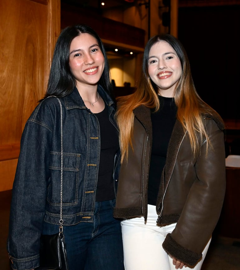 Dos mujeres sonriendo, una con abrigo de mezclilla oscuro y otra con chaqueta marrón, en un ambiente con cortinas rojas y tenue iluminación.