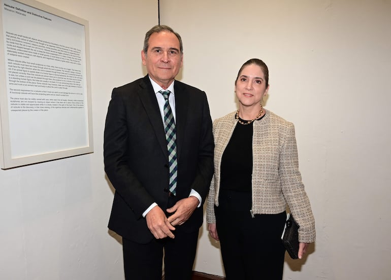 Hombre en traje oscuro con corbata a rayas y mujer en abrigo claro junto a una pared con texto de la exposición 'Netsuke'.
