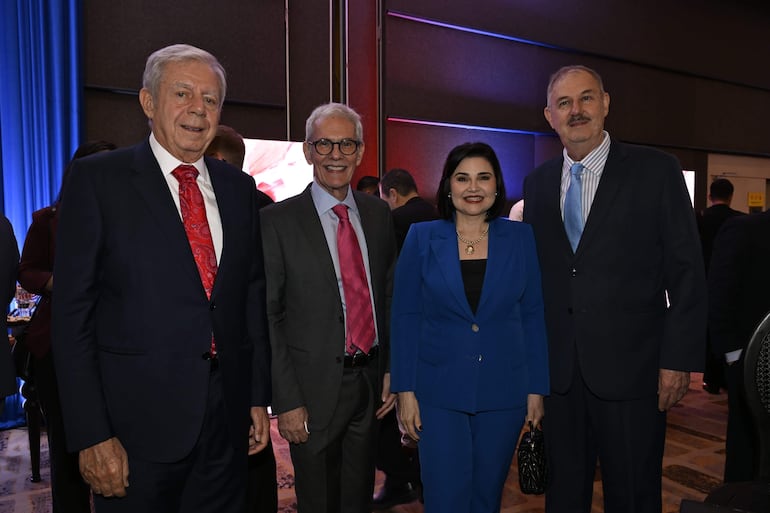 Dos hombres y dos mujeres sonrientes en un ambiente decorado con luces azules y rojas, visten trajes elegantes y mantienen una conversación.
