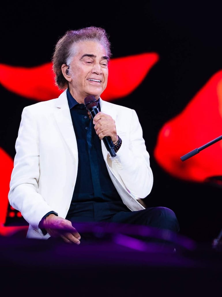 Hombre de cabello rizado y canoso, con chaqueta blanca y camisa negra, sostiene un micrófono en el escenario iluminado.