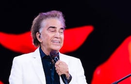 Hombre de cabello rizado y canoso, con chaqueta blanca y camisa negra, sostiene un micrófono en el escenario iluminado.
