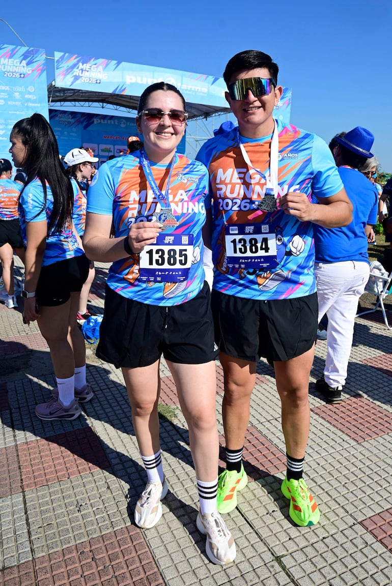 Mujer y hombre sonrientes sostienen medallas con camisetas deportivas azul celeste en evento de carrera al aire libre.