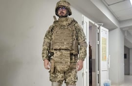 Hombre en uniforme militar de camuflaje, con casco y gafas, de pie en un pasillo interior con paredes claras.
