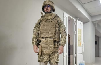 Hombre en uniforme militar de camuflaje, con casco y gafas, de pie en un pasillo interior con paredes claras.