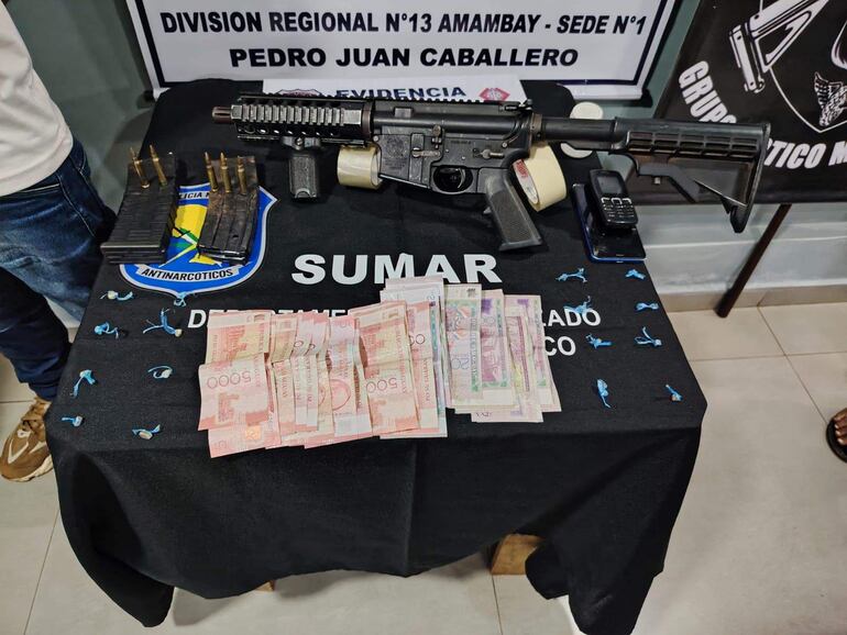 Rifle de asalto y evidencias sobre mesa negra en ambiente antinarcóticos, con cartel de 'Pedro Juan Caballero' al fondo.