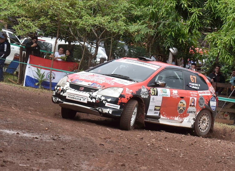 Auto Honda número 67 en acción, personas observando y agitando una bandera de Paraguay en un entorno natural.