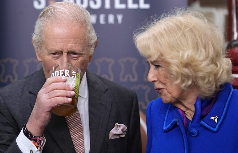 La reina Camilla reacciona mientras el rey Carlos III prueba una pinta de Proper Job IPA 0.5% de la cervecería St Austell que él mismo sirvió, durante una visita al Mercado Central en St Austell, en el suroeste de Inglaterra. (Aaron Chown / POOL / AFP)