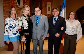 Hombres y mujeres en atuendos formales, sonriendo en un evento social en una sala con ladrillos expuestos y banderas.