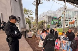Oficial de seguridad de pie, mientras una mujer vestida de negro habla con niños en un entorno urbano.