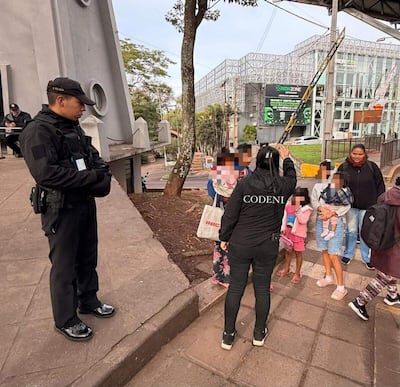 Oficial de seguridad de pie, mientras una mujer vestida de negro habla con niños en un entorno urbano.