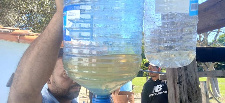 Hombre con botella de agua amarilla y otro observando, rodeados de césped y árboles en un espacio natural.