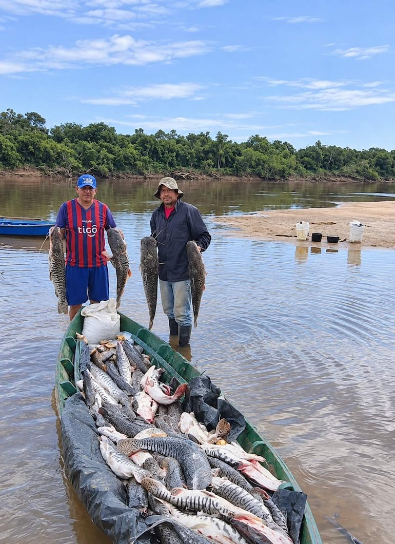 Hombres en el río sosteniendo grandes peces, uno con camiseta a rayas rojas y azules y gorra azul, otro con camiseta negra y sombrero beige.