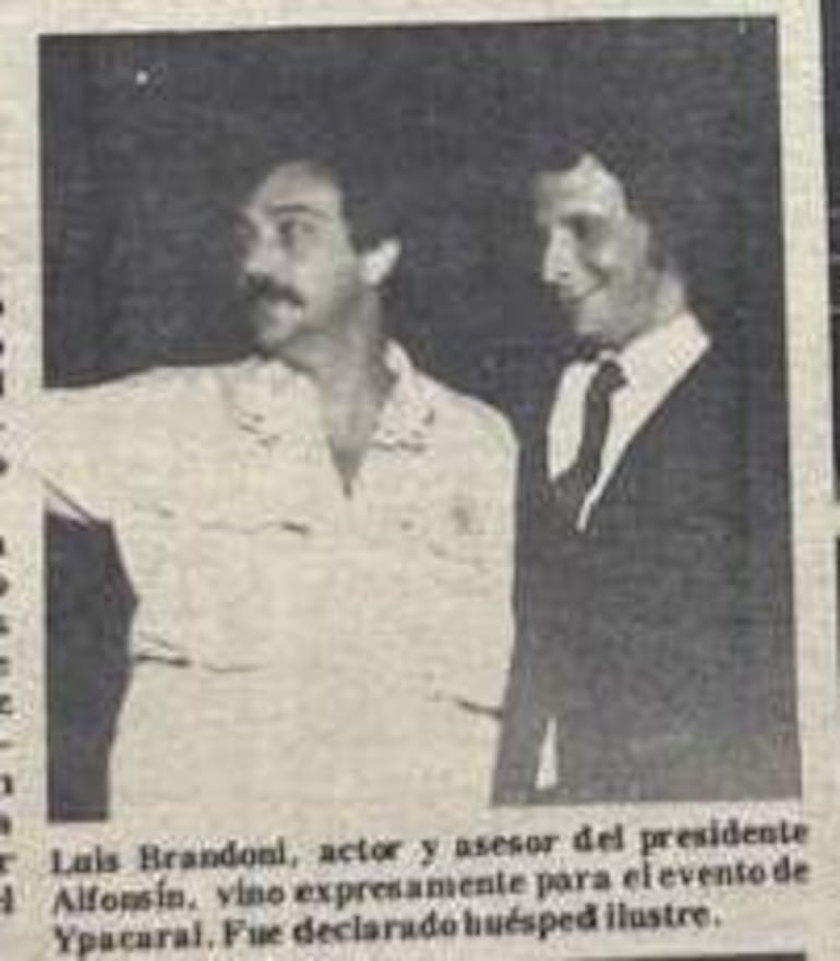 Luis Brandoni gesticula con entusiasmo en un ambiente informal, mientras otro hombre en traje lo escucha atentamente.