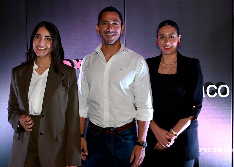 Mujer con blazer marrón y blusa blanca sonríe a la izquierda; hombre con camisa blanca y jeans en el centro; mujer en atuendo negro a la derecha, todos en un evento formal.