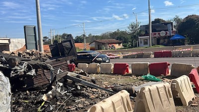 Escena urbana con restos carbonizados de estructura y un coche negro estacionado frente a barricada.