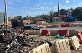 Escena urbana con restos carbonizados de estructura y un coche negro estacionado frente a barricada.
