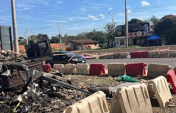 Escena urbana con restos carbonizados de estructura y un coche negro estacionado frente a barricada.
