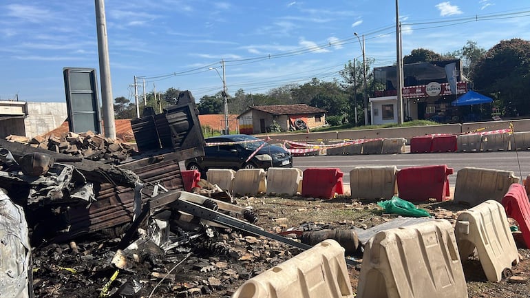 Escena urbana con restos carbonizados de estructura y un coche negro estacionado frente a barricada.