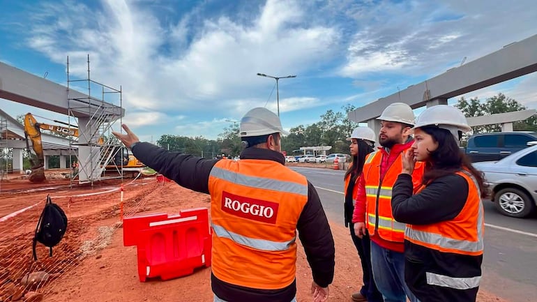 Cuatro trabajadores en chalecos anaranjados discuten sobre la obra en construcción, con excavation y estructuras metálicas de fondo.